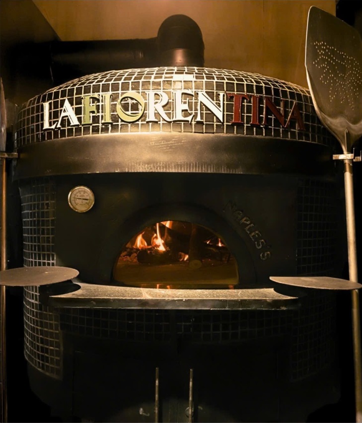 Chiếc lò nướng pizza bằng gạch kiểu Napoli truyền thống, khảm dòng chữ 'LA FIORENTINA', đang cháy lửa với xẻng pizza bên cạnh tại nhà hàng Ý La Fiorentina Tây Hồ